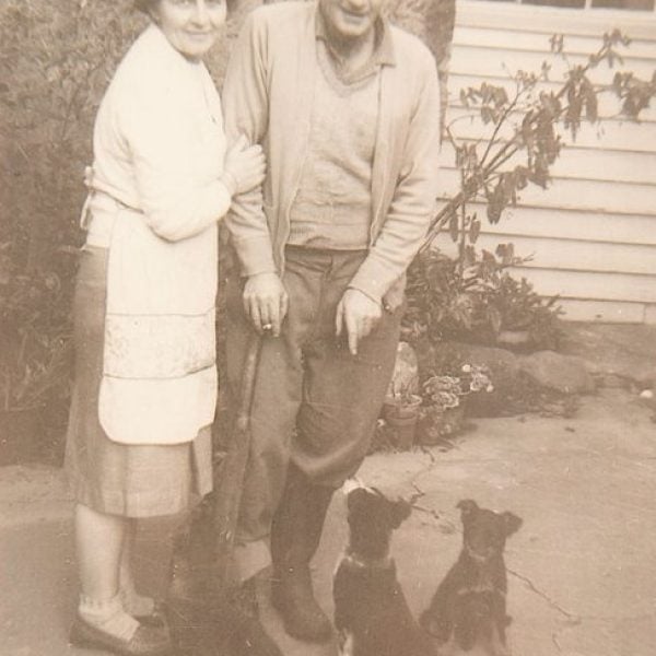 Old photo of two people standing outside a building with two small dogs