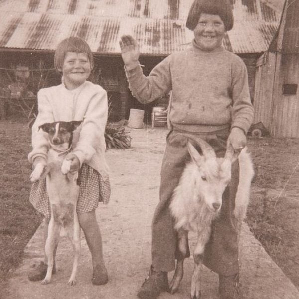 Old photo of two kids with a goat and a dog, a house is in the background