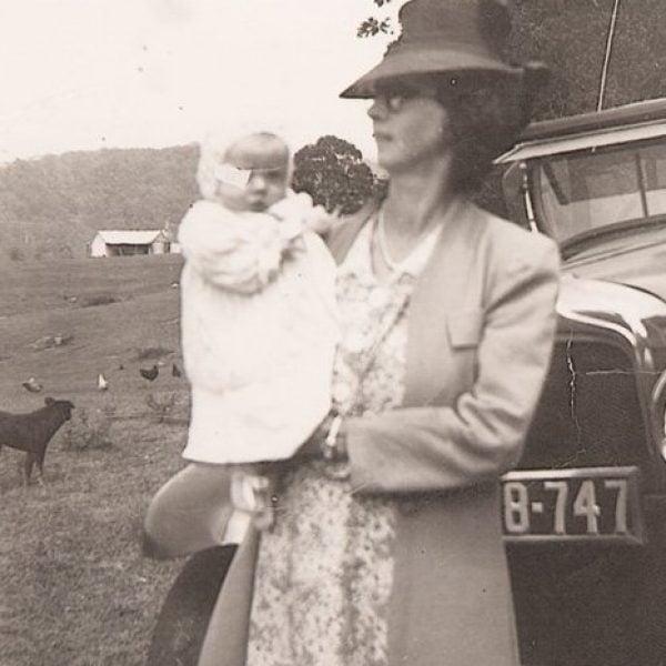 Old photo of a woman holding a baby standing next to a car. Dog in the background