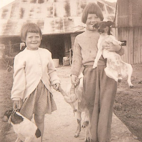 Old photo of two kids with a goat and a dog, a house is in the background