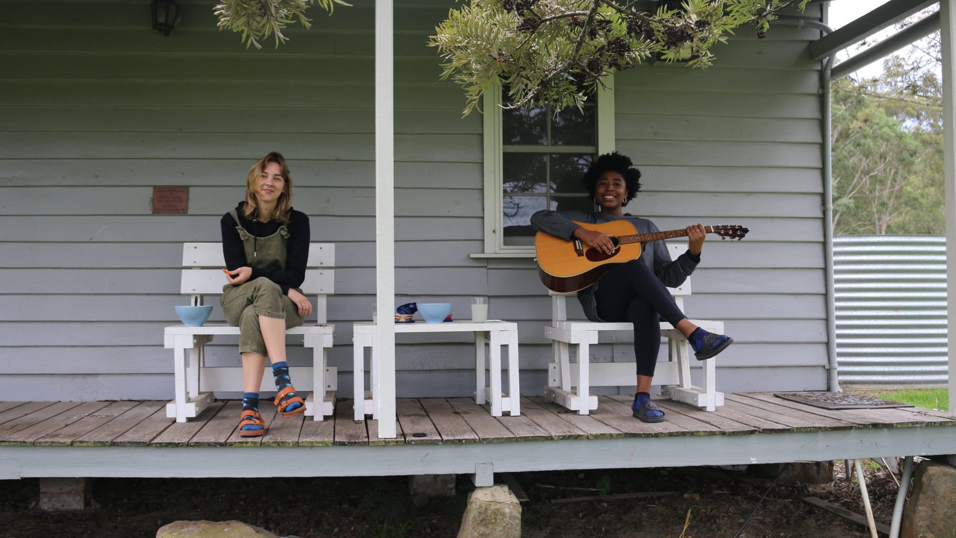 Two people sitting on the verandah of a grey cottage. The 1 person is holding a guitar