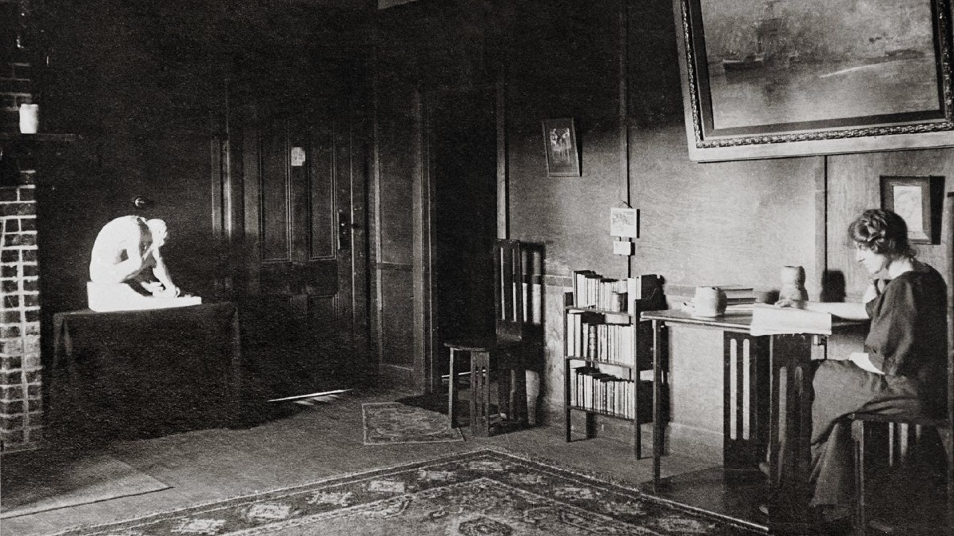 Old photo of a room with sculptures and paintings. A woman sits writing at a desk