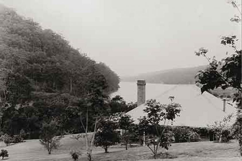 Black and white photo of an old house and a river and mountains in the background