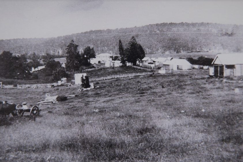 Black and white photo of farm buildings in a field surrounded by mountains