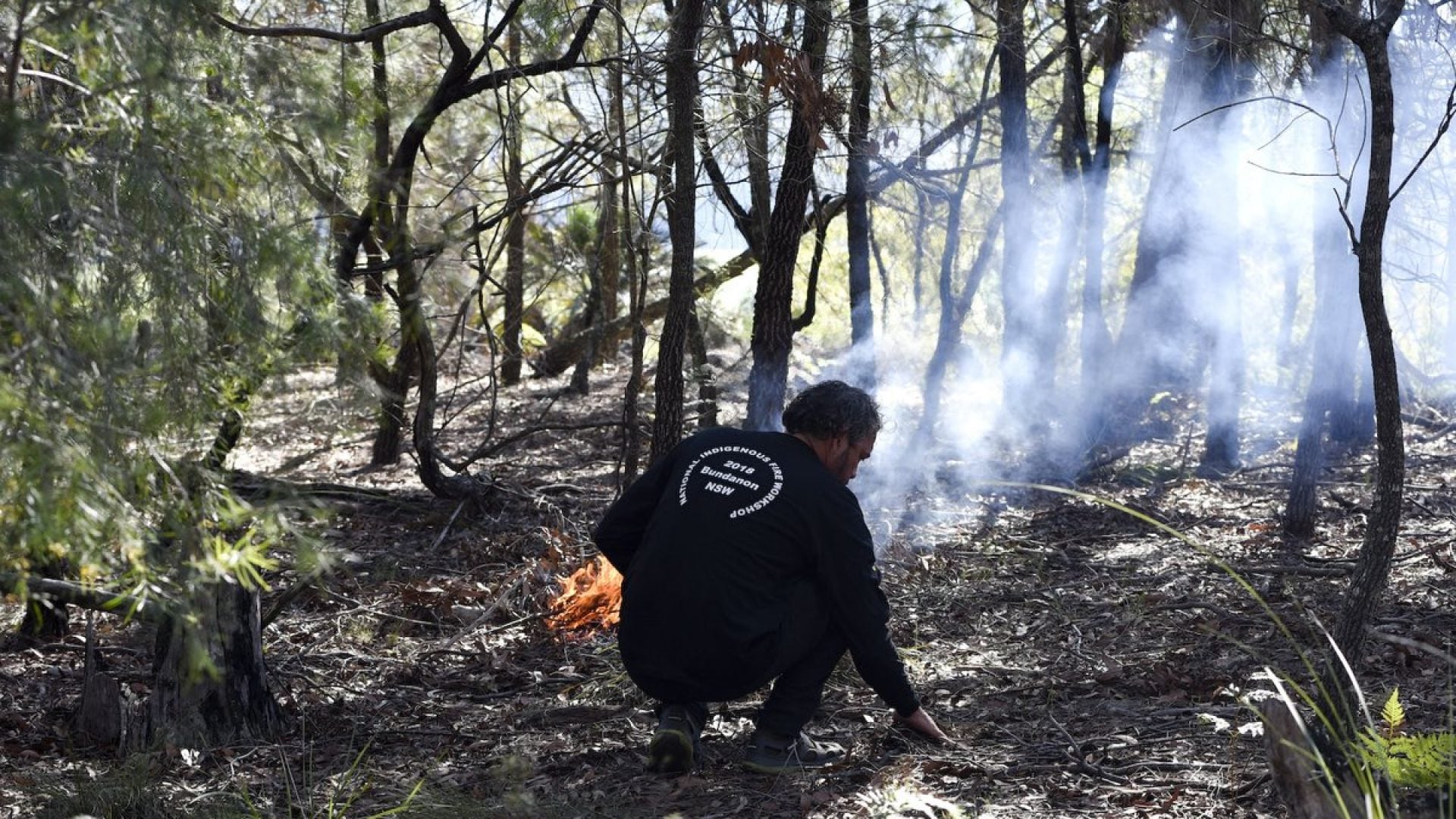 Person kneeling in a forest landscape with a small fire burning beside them