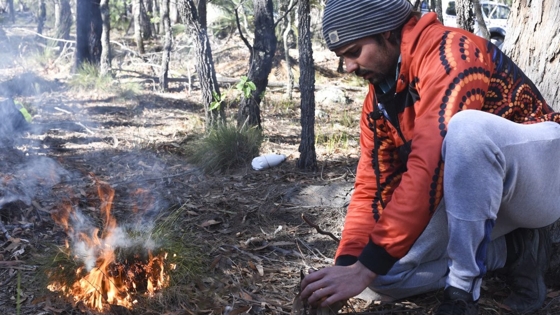 Person kneeling in a forest landscape with a small fire burning beside them