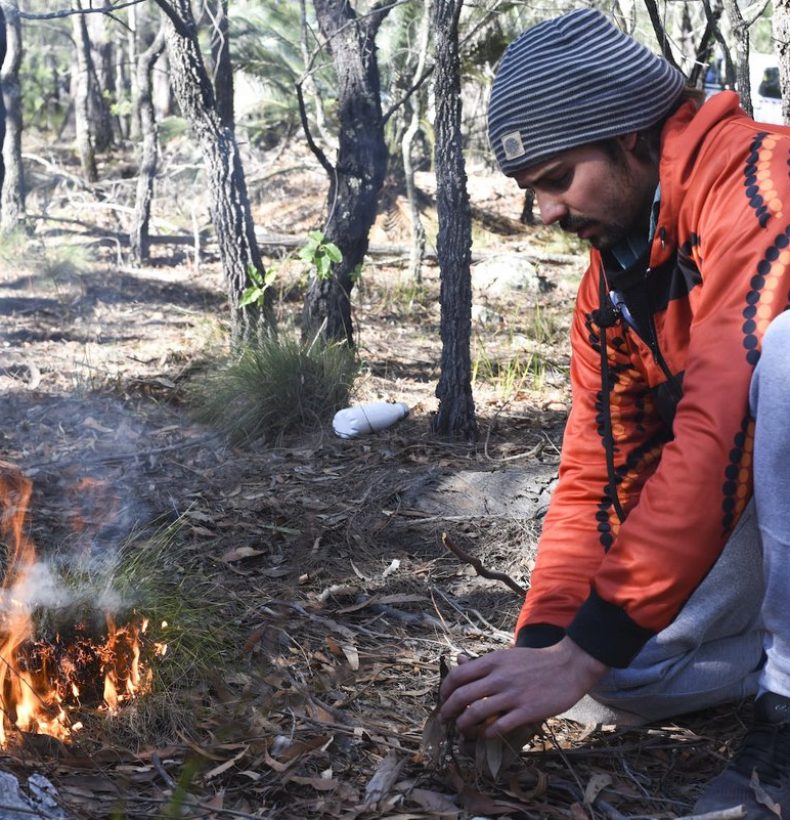 Person kneeling in a forest landscape with a small fire burning beside them