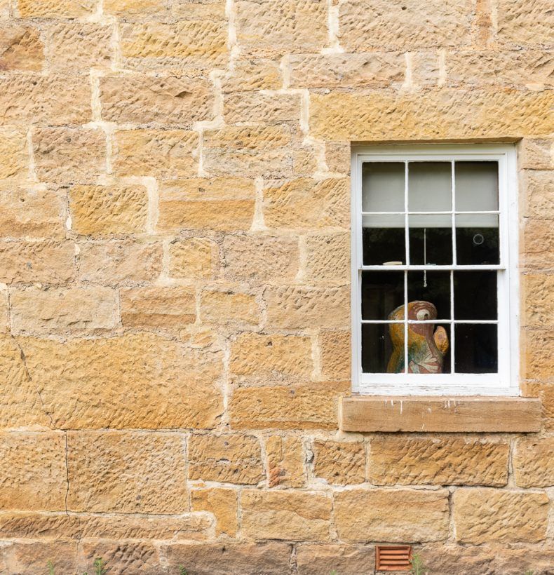 The sandstone wall of a building. A window in the centre with a small sculpture visible inside.