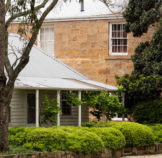 A wooden white building stands infront of a tall sandstone building. All surrounded by gardens and trees