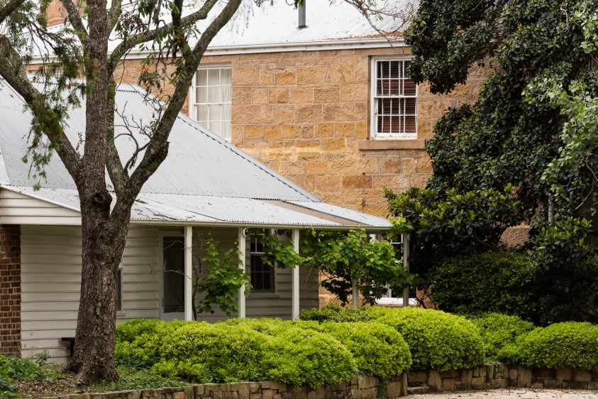 A wooden white building stands infront of a tall sandstone building. All surrounded by gardens and trees