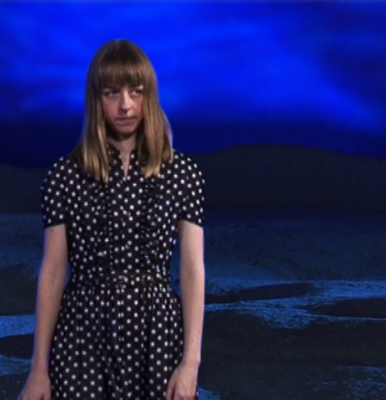 Person with shoulder length hair stands in a room with sand on the ground, the room is illuminated with blue lighting