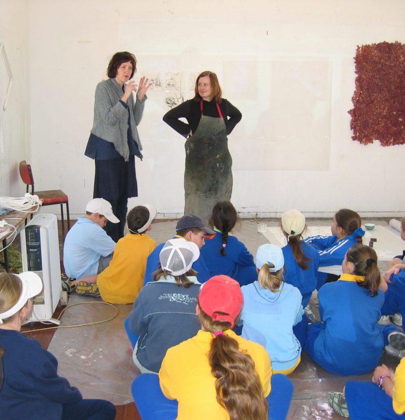 Two adults standing in front of 16 school children. There are abstract drawings on the wall.