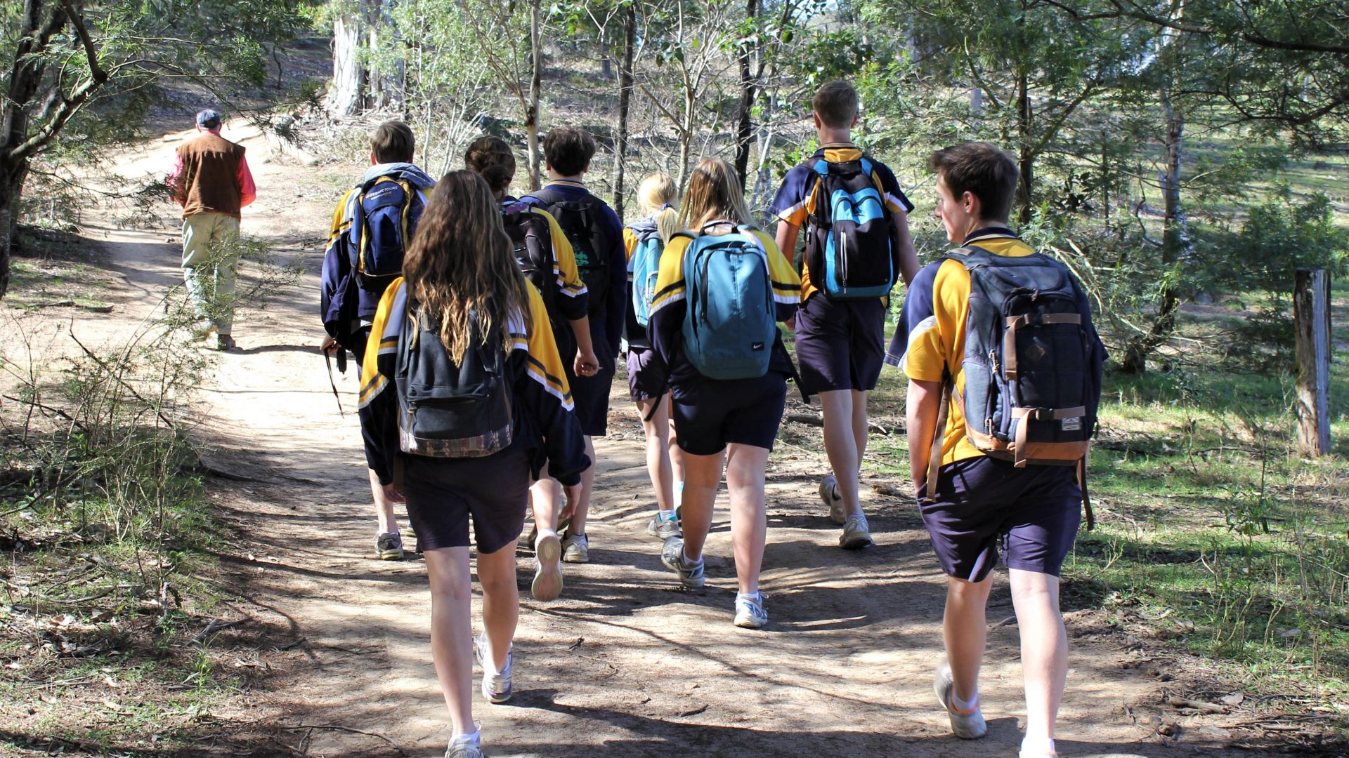 A group of students with backpacks walking along a dirt road surrounded by bush land