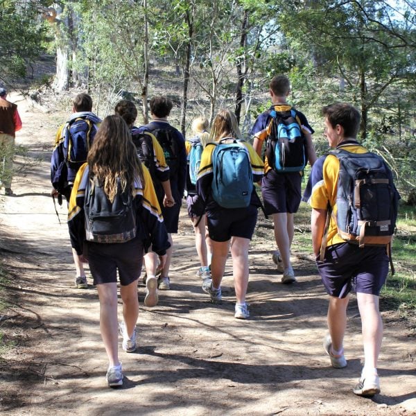 A group of students with backpacks walking along a dirt road surrounded by bush land