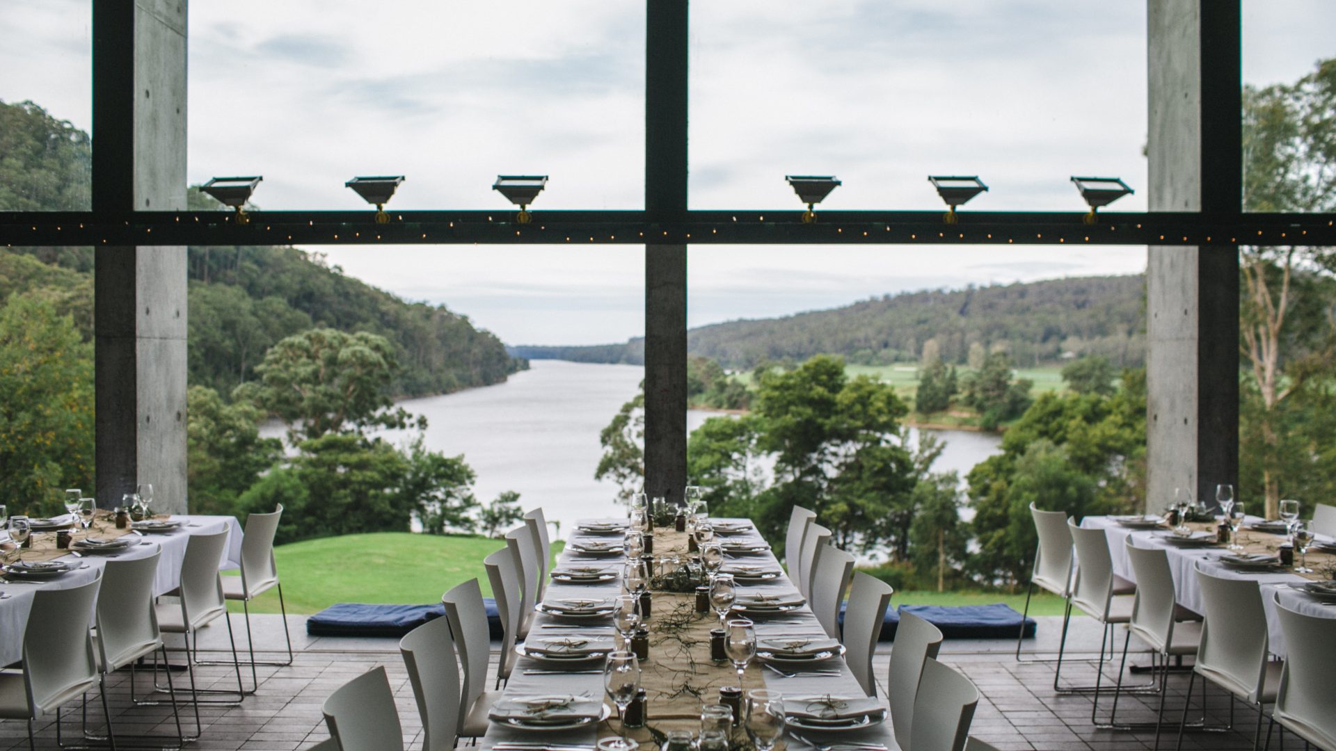 Rows of set dining tables in a room with a large window overlooking a river and mountains.