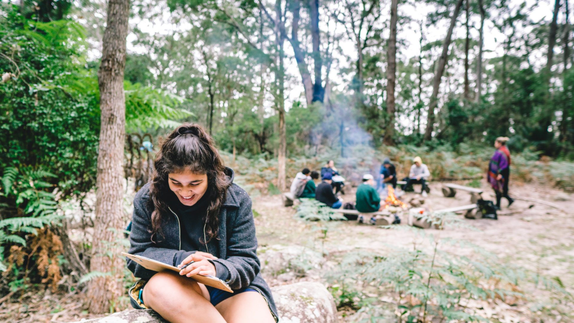 A girl sitting on a rock reading a book. Behind her a group sits around a camp fire