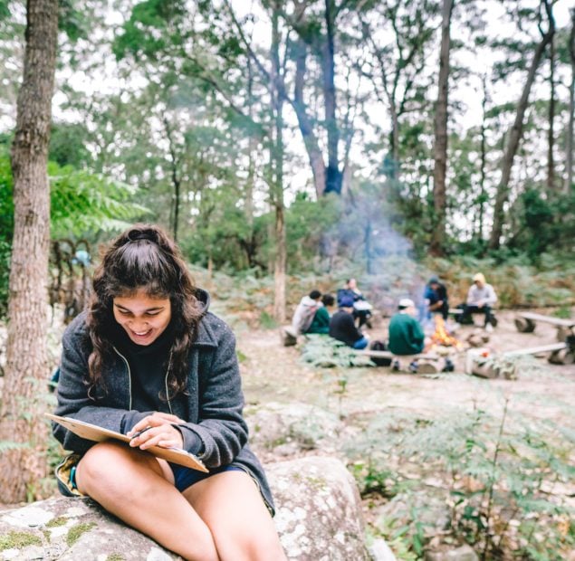 A girl sitting on a rock reading a book. Behind her a group sits around a camp fire