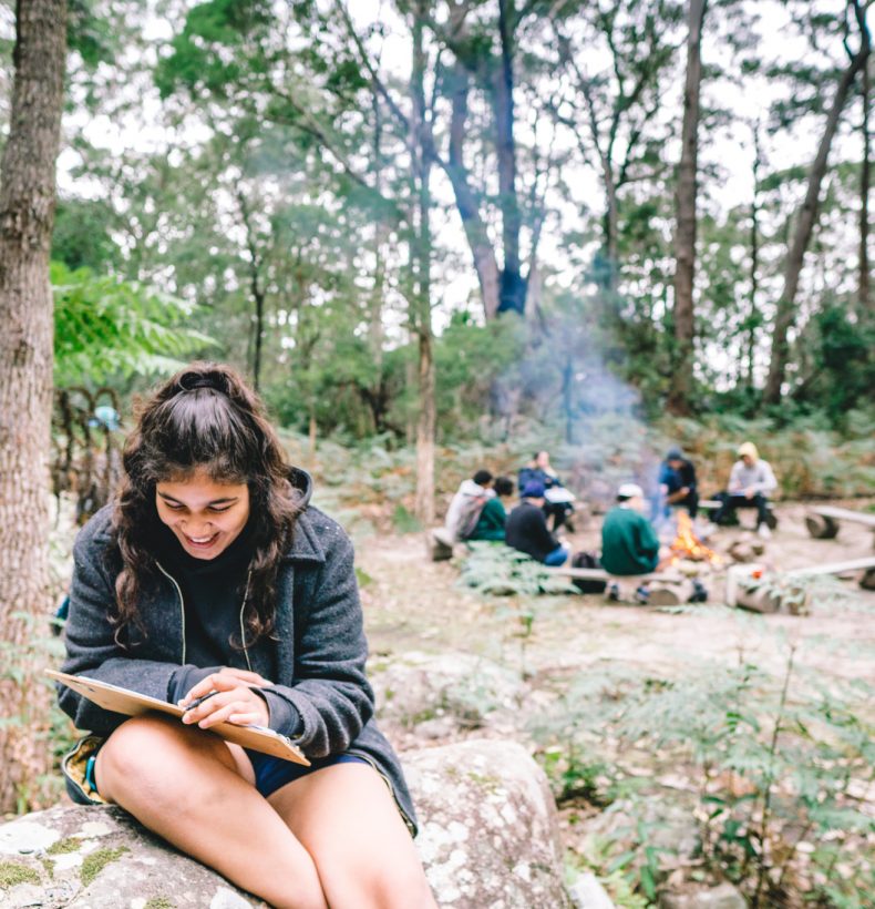 A girl sitting on a rock reading a book. Behind her a group sits around a camp fire