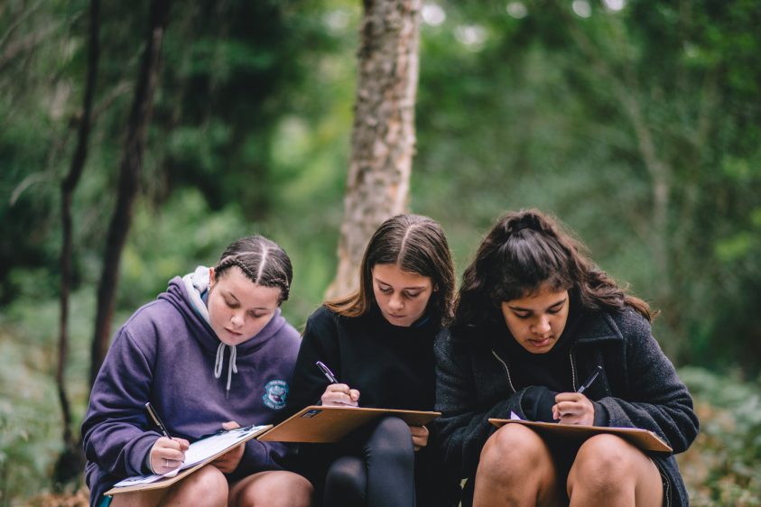 Three girls with long hair sit together writing on pieces of paper. They are surrounded by bush land