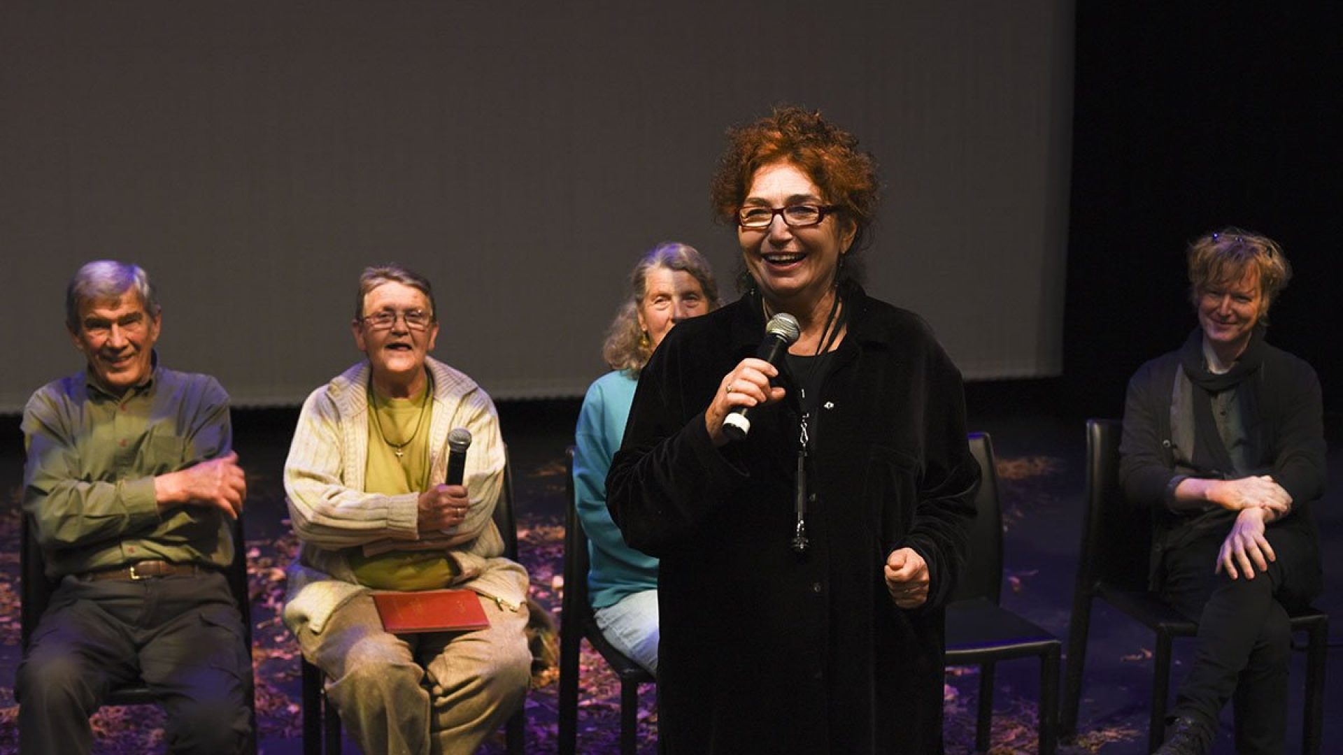 A person standing with a microphone. 4 people behind them are sitting on chairs