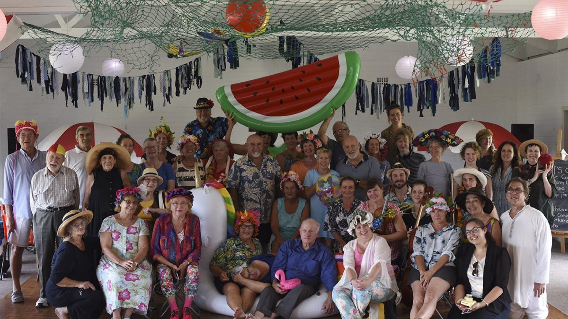 A group of 30 people wearing bright flower head dresses holding a large inflatable watermelon