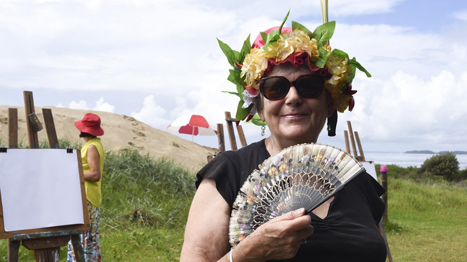 Person standing on the beach wearing a floral head dress and holding a fan in front of them