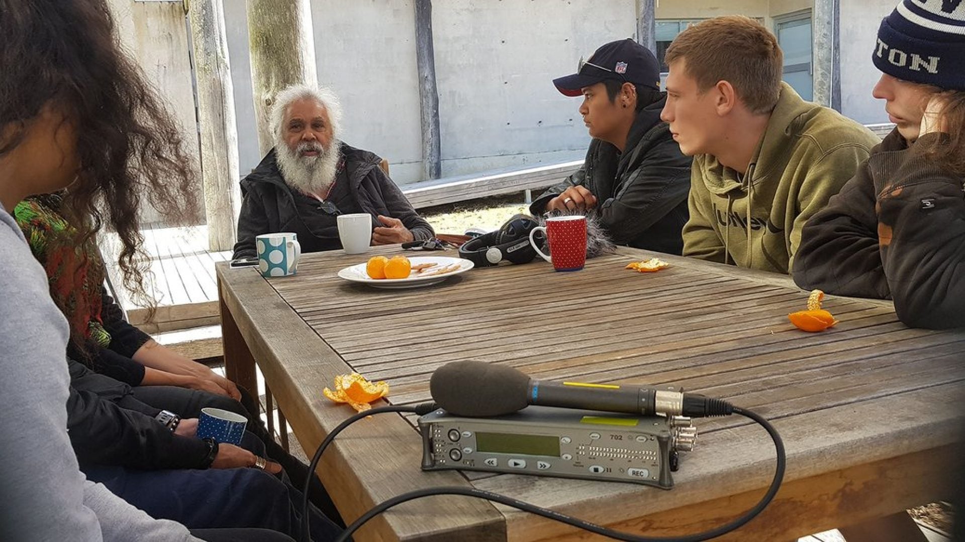 6 young people sitting at a wooden table looking at a person with white hair and a white beard