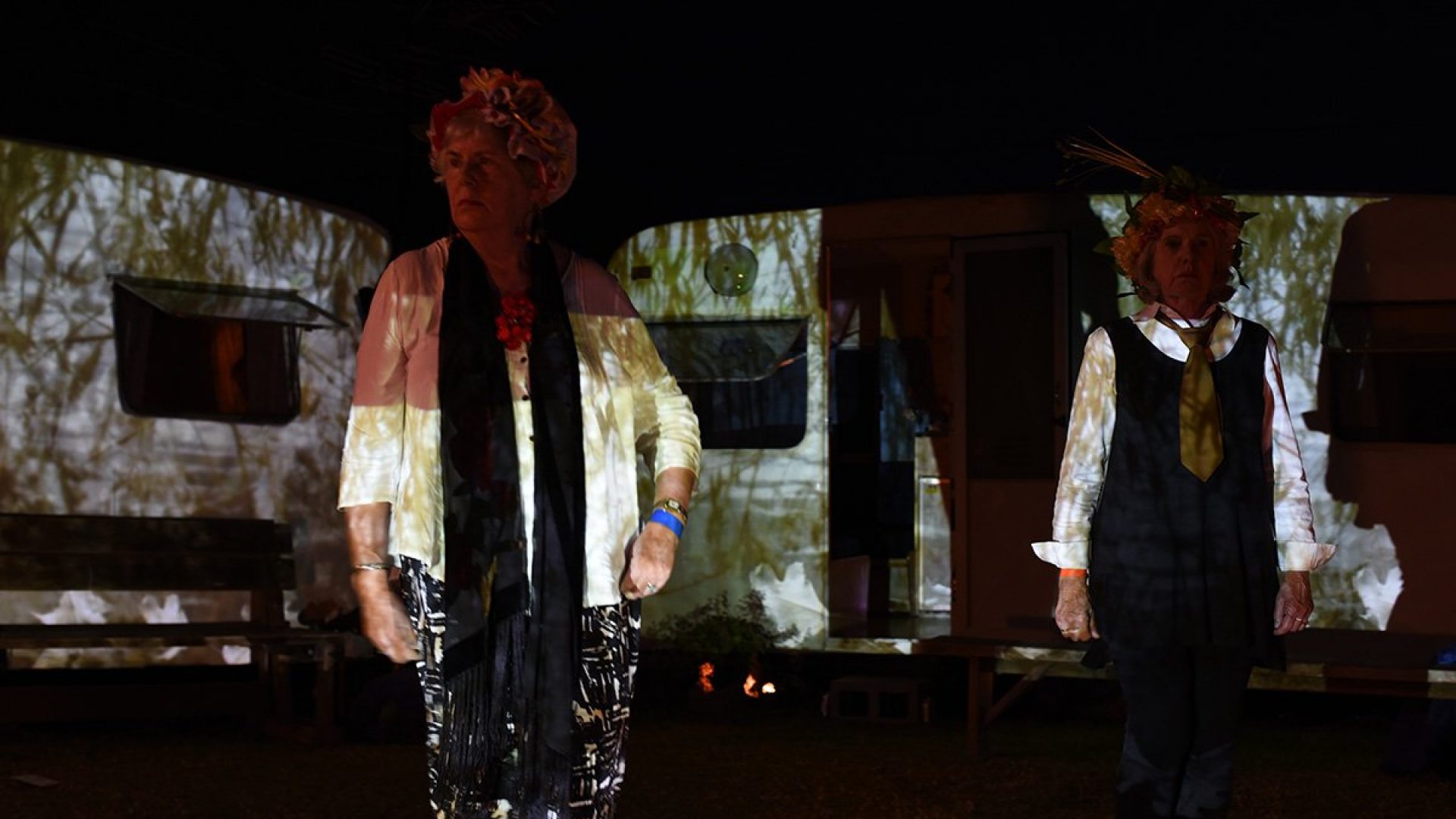 2 people sitting in front of two old caravans that have trees and birds projected on them