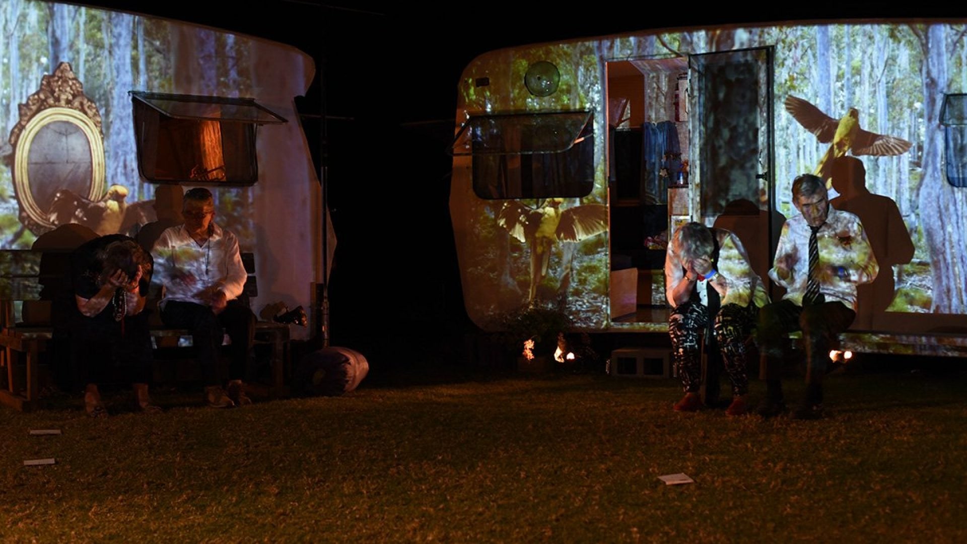 People sitting in front of two old caravans that have trees and birds projected on them