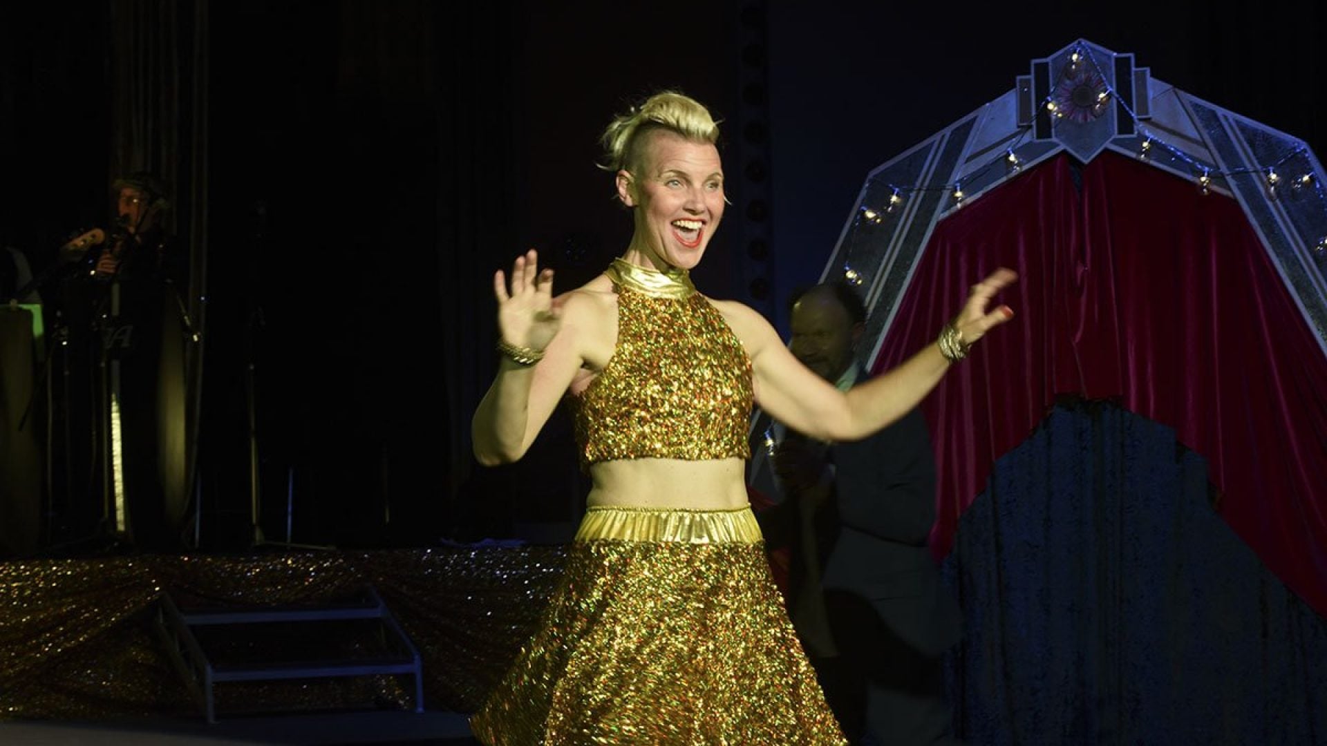 A person in a gold dresses stands with their arms up and mouth open