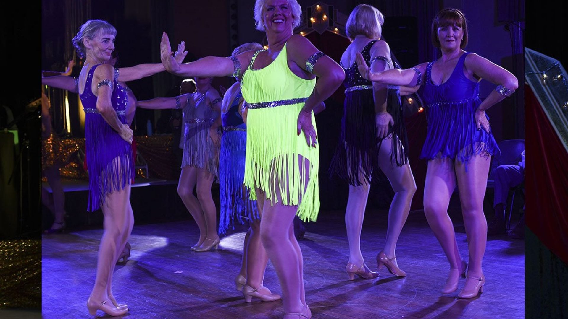 6 women standing on stage wearing short blue and yellow flapper dresses