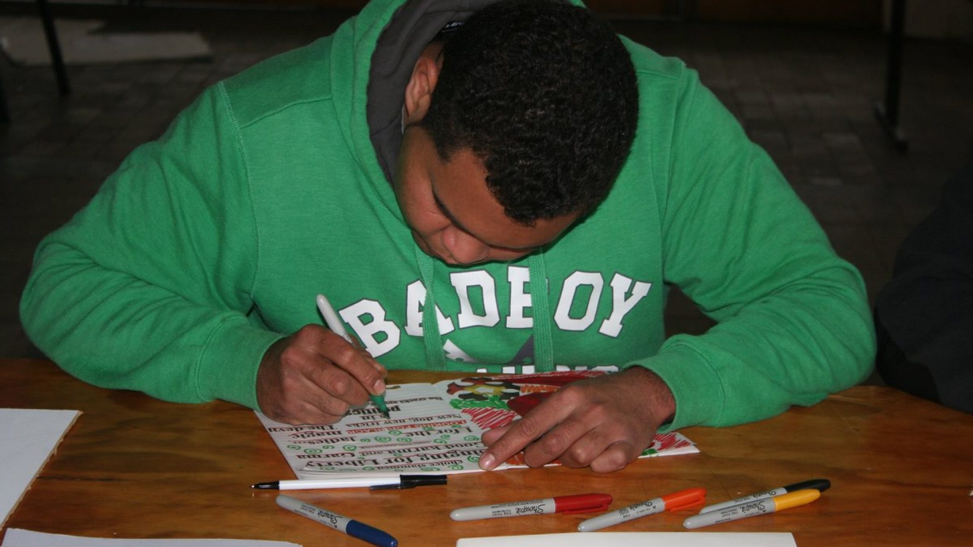 Person with short dark hair drawing a poster. They are wearing a green hooded jumper