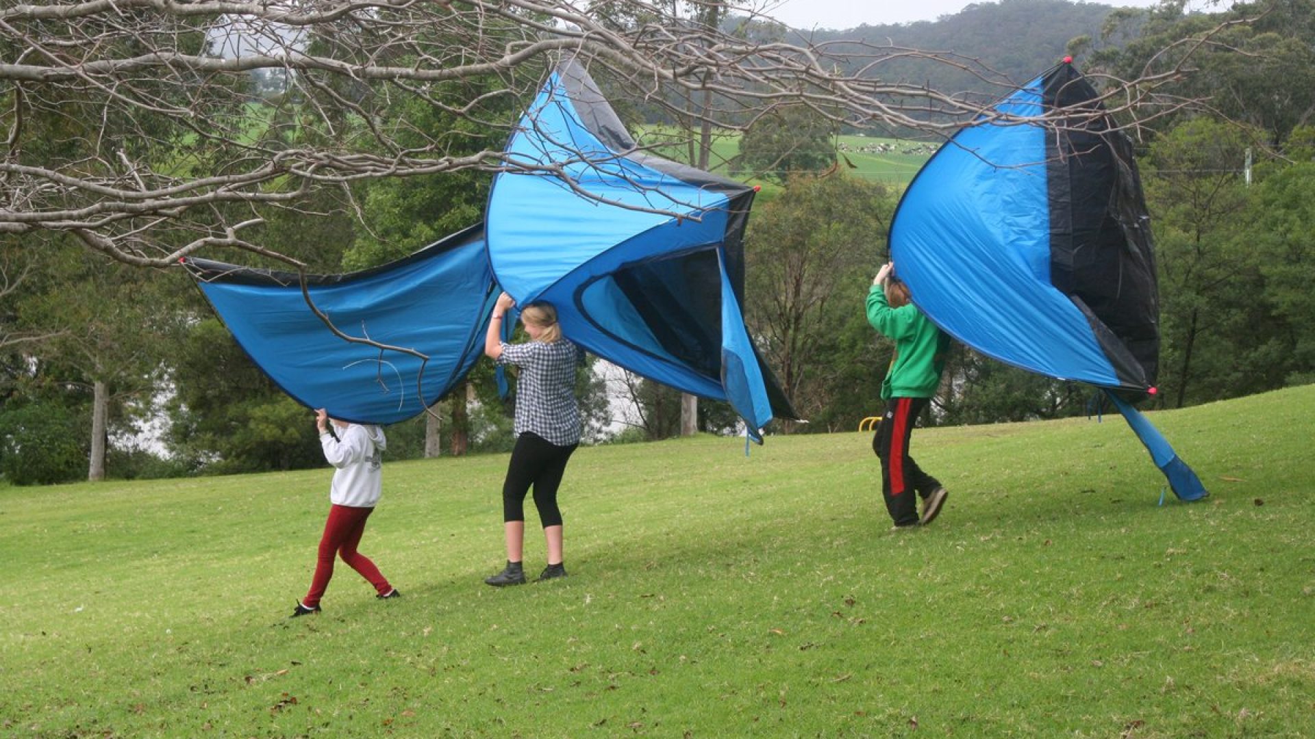 3 people holding a blue tent each walk down a grassy hill towards a river