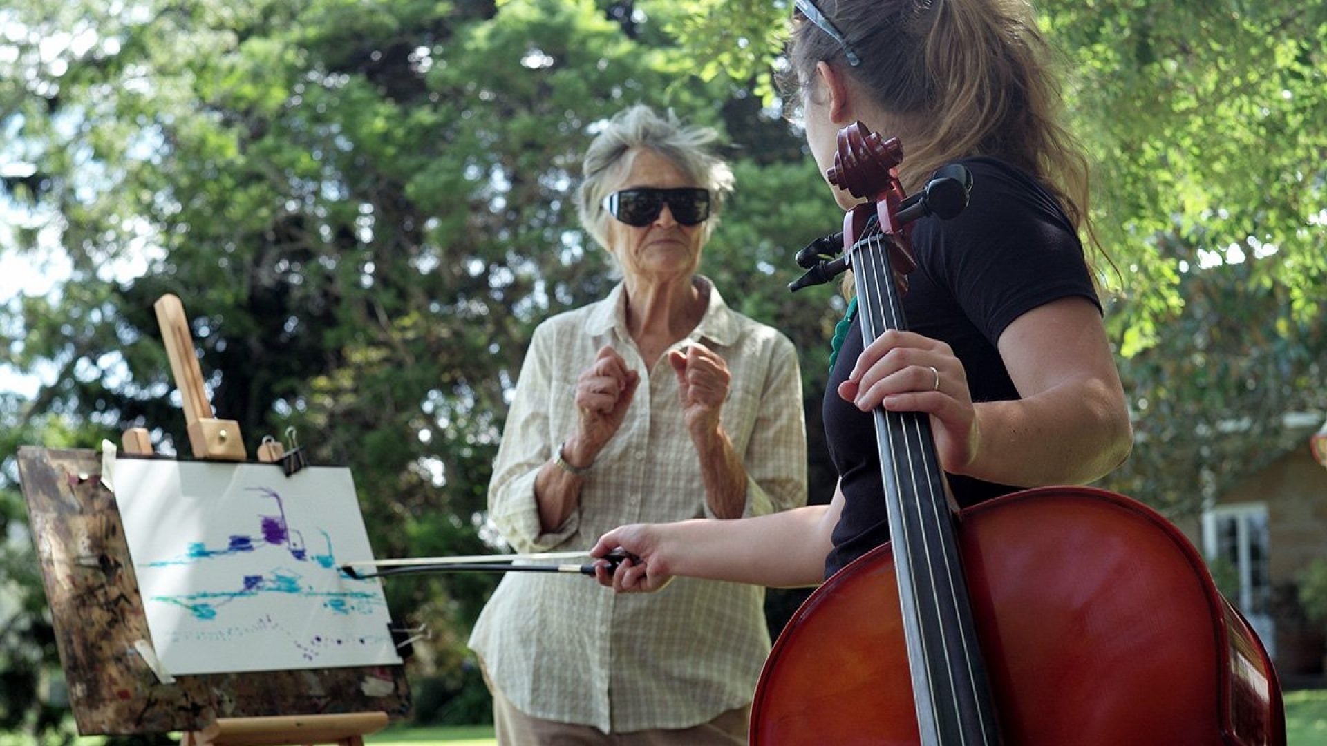 A person holding a cello talks to someone drawing on an easel