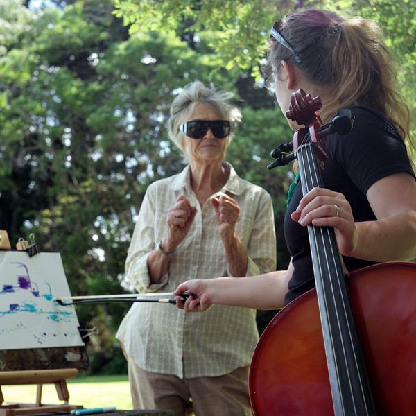 A person holding a cello talks to someone drawing on an easel