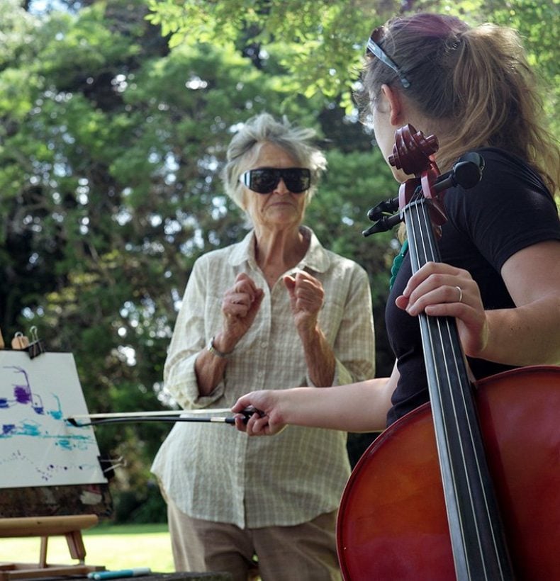 A person holding a cello talks to someone drawing on an easel