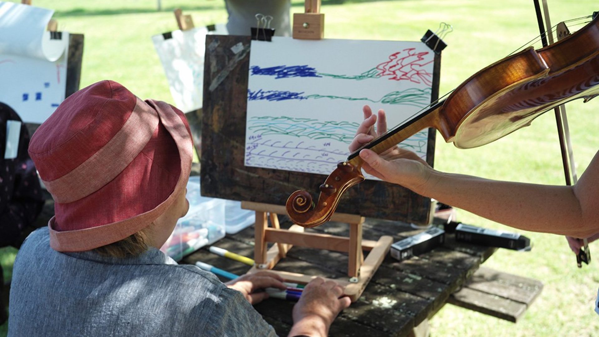 A person sitting a wooden bench drawing while a person stands beside them playing a violin