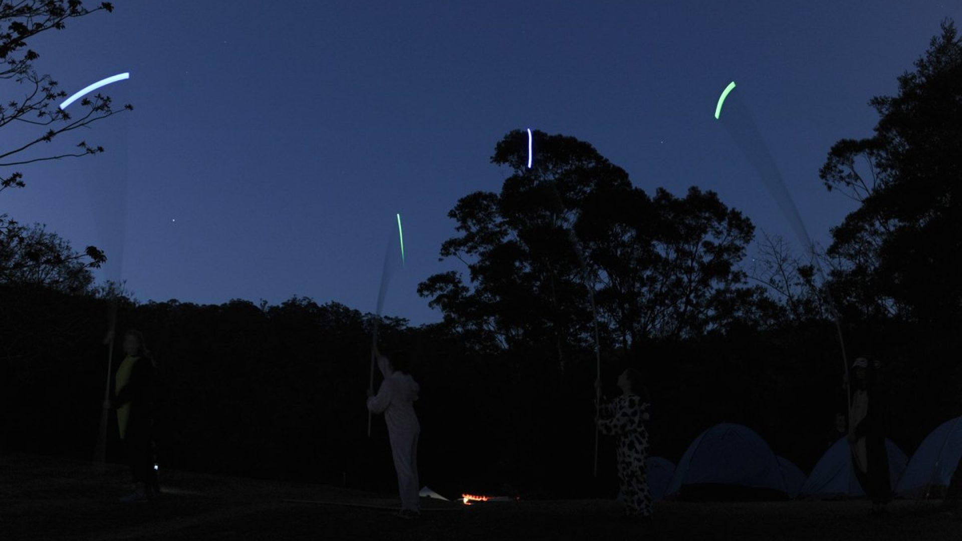 4 people stand in a dark landscape holding tall rods with lights on the ends of them