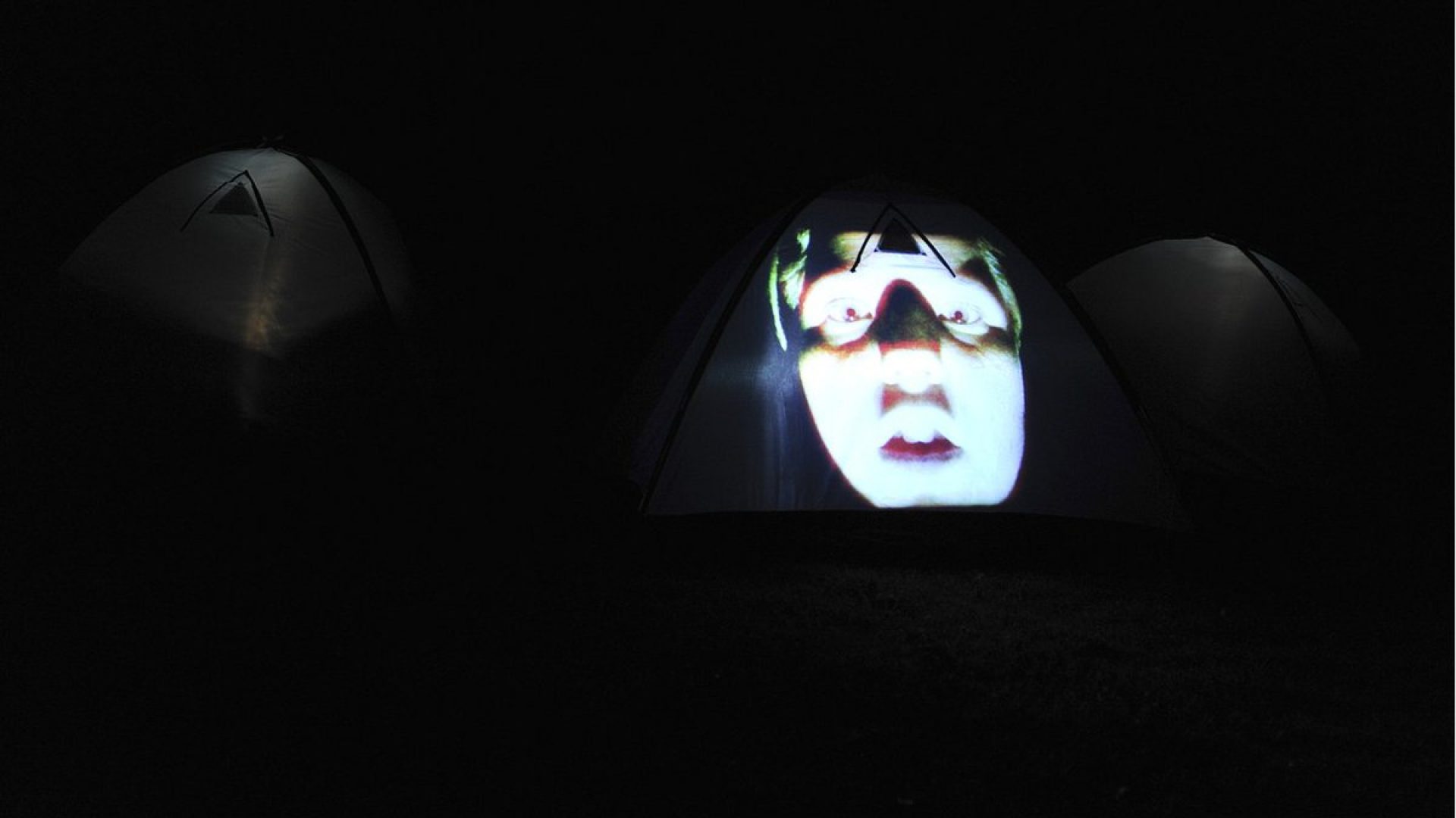 3 tents in a dark landscape with a face image projected on the side of one tent