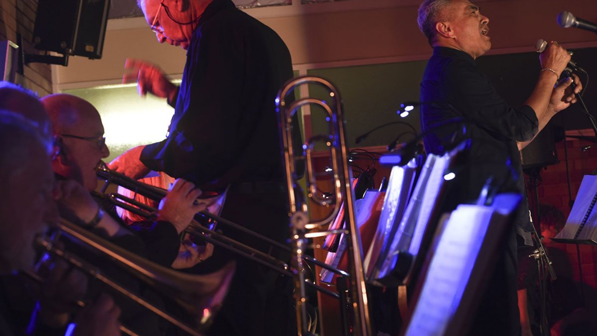 4 people playing brass instruments, a person is beside them singing into a microphone