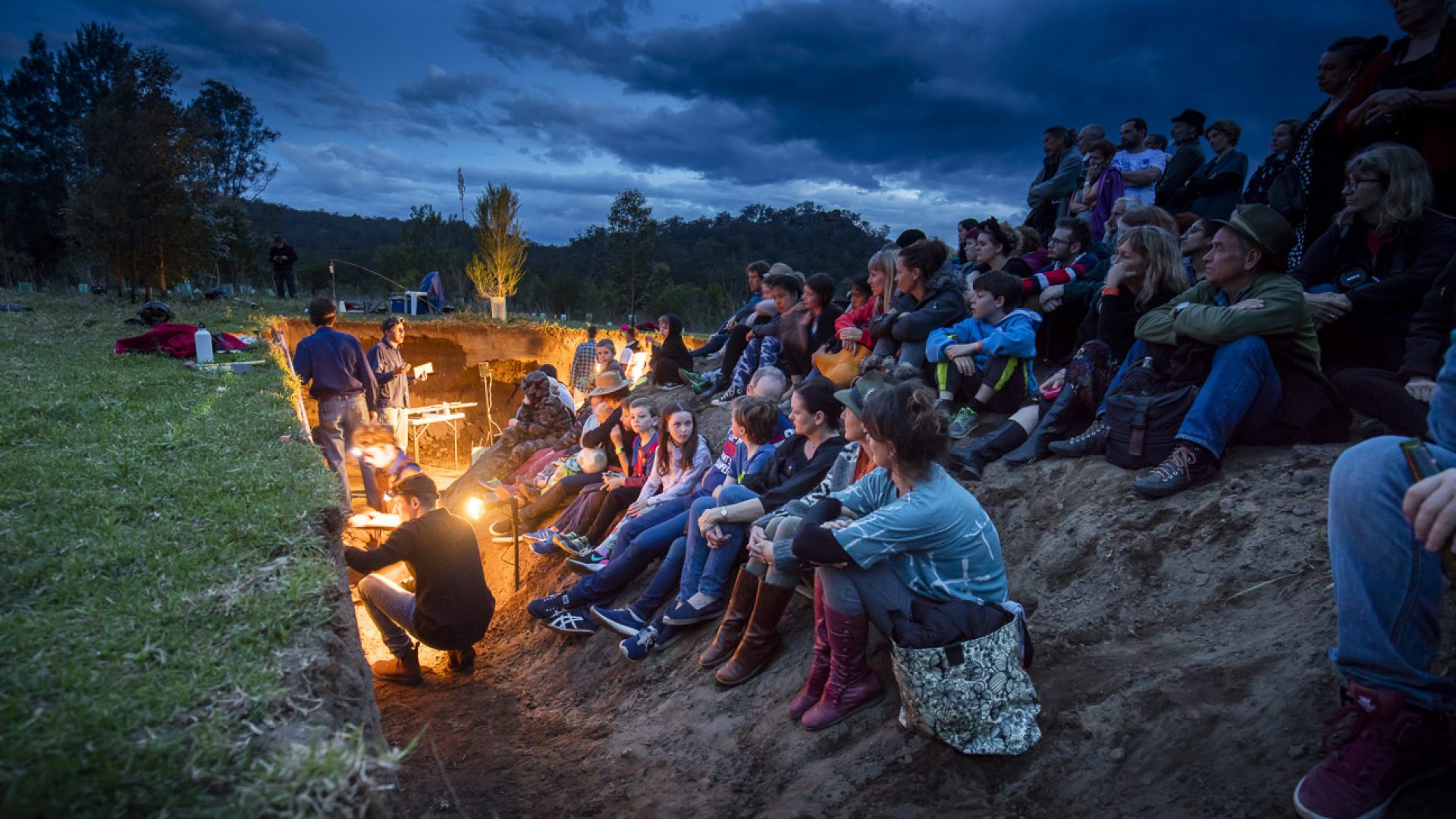 A group of 50 people sit around an excavated hold in the ground