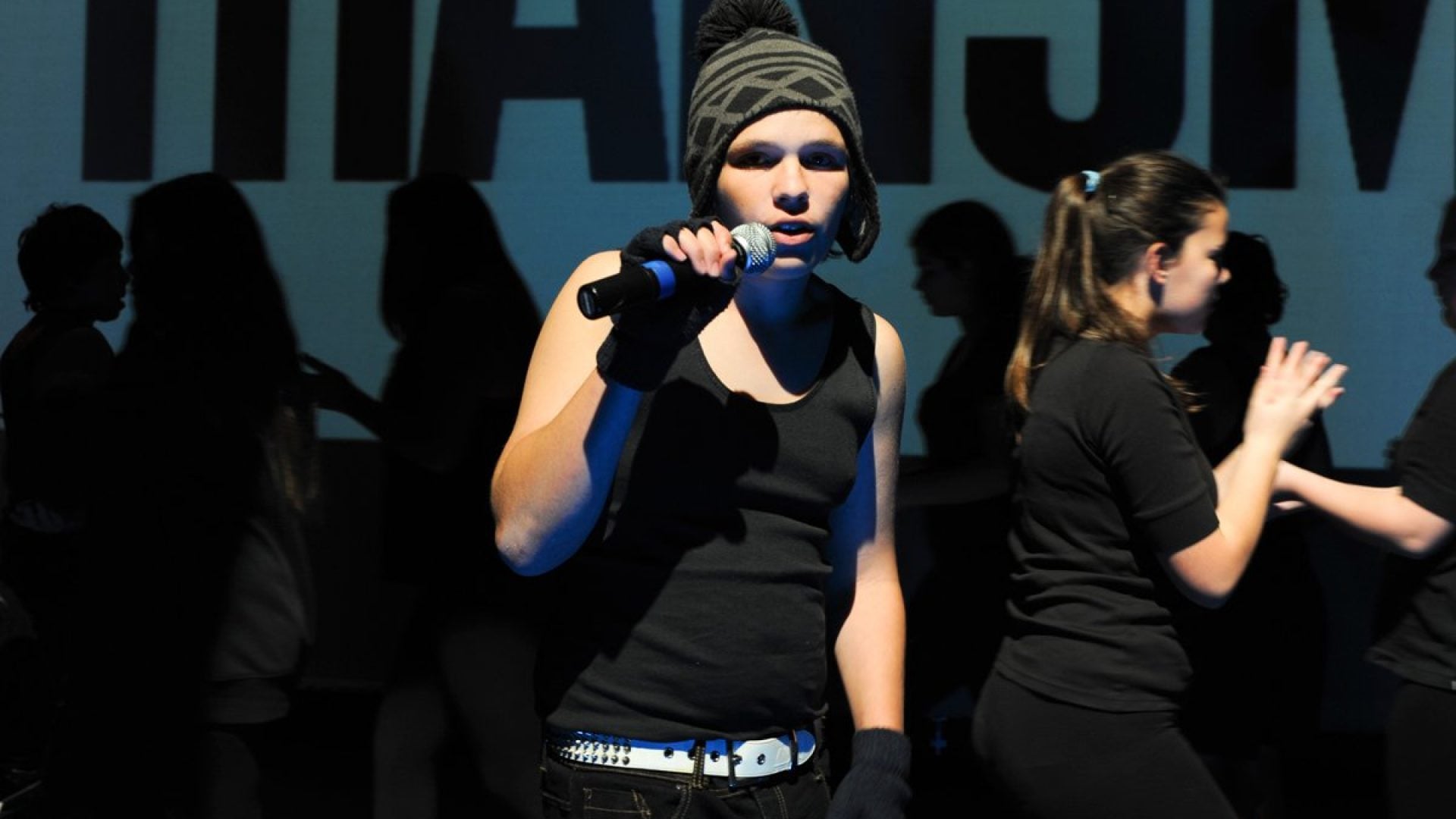 Young person holding a microphone on a stage with dancers behind them