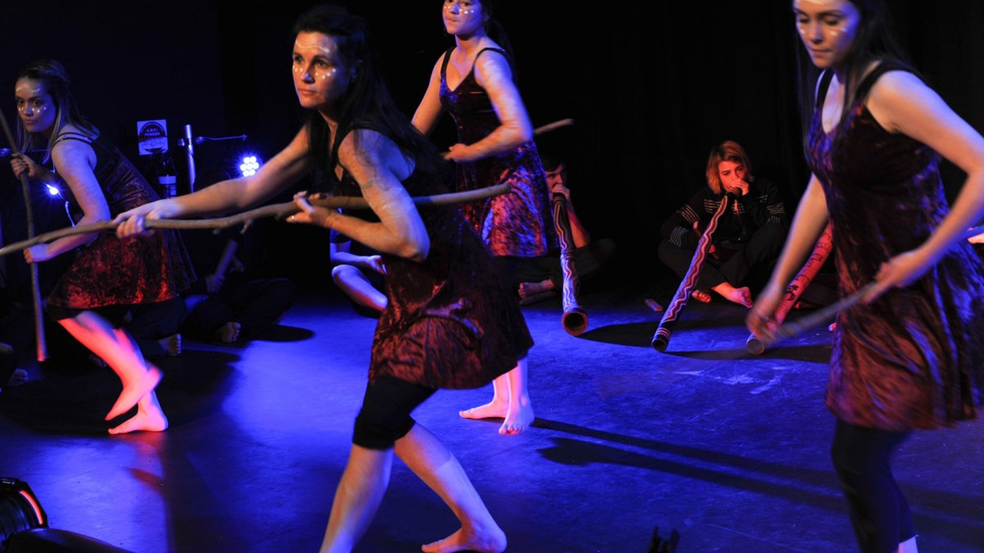 4 people dancing. Didgeridoo players sitting on the ground behind them