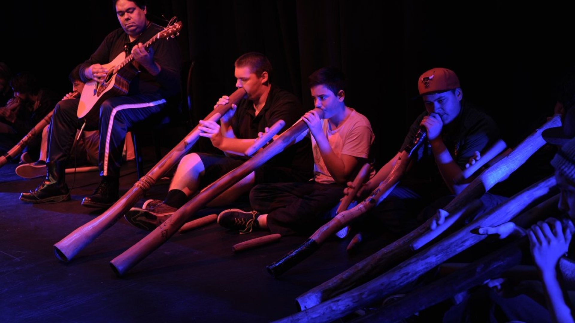 8 people sitting playing the didgeridoo. A person sits on a seat playing the guitar in the middle of them