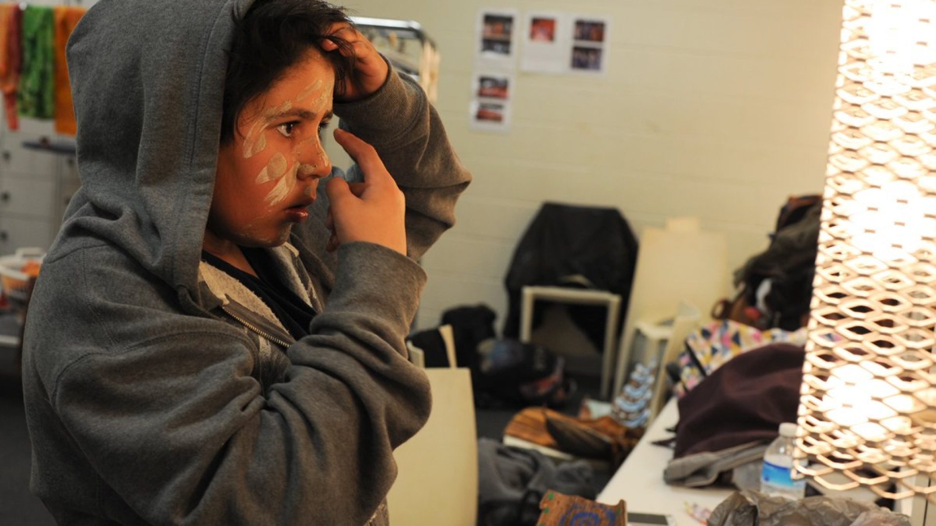 Young boy in a hoodie looking into a mirror, putting paint on his face