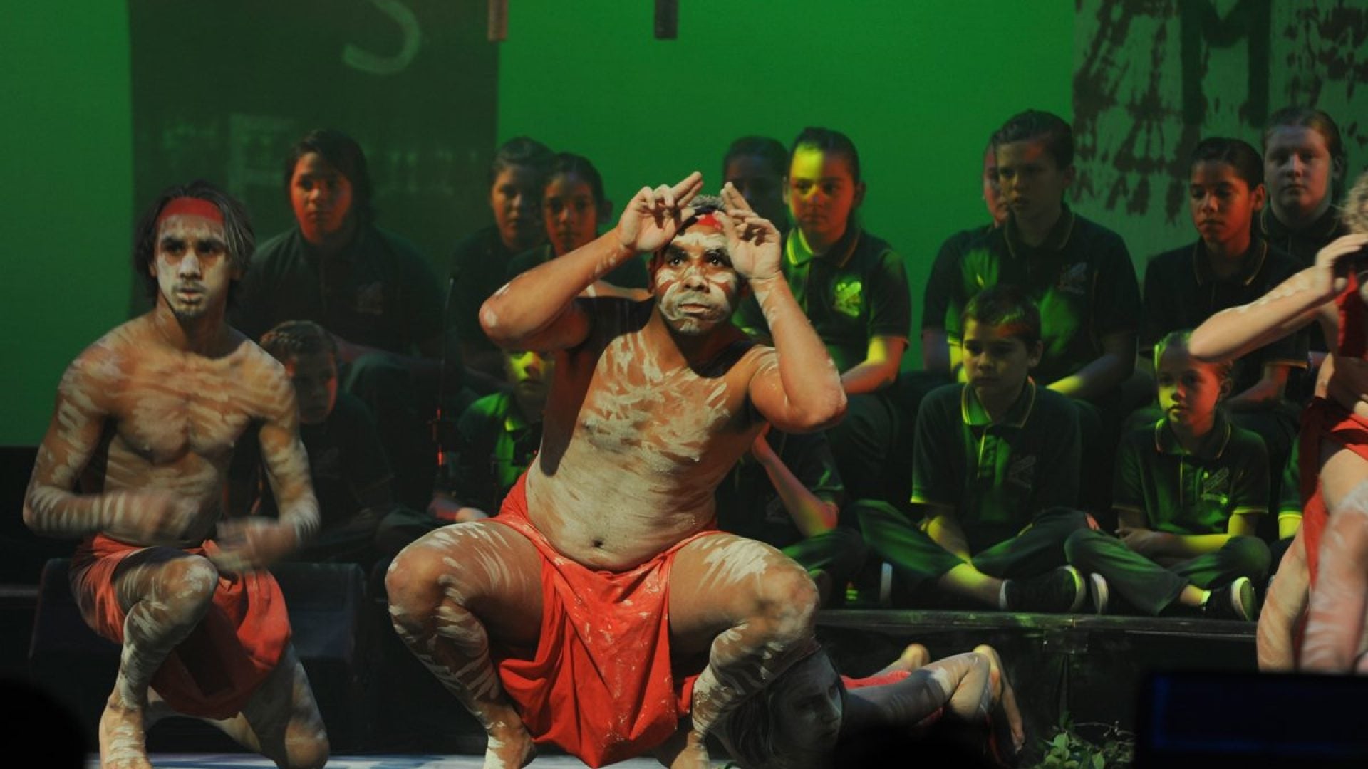 3 traditional aboriginal dancers. A choir of children sitting on stage behind them