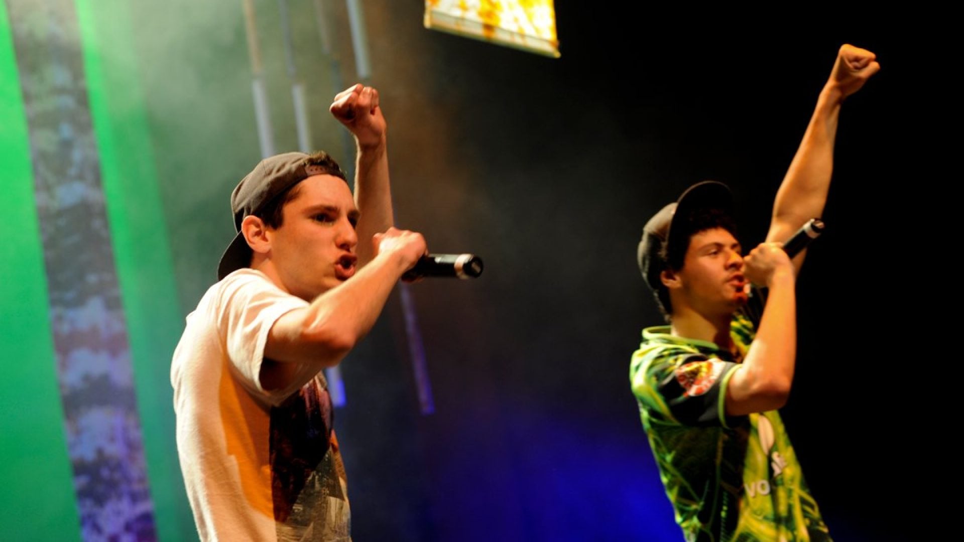 Two people standing on stage singing into microphones with their left fists in the air