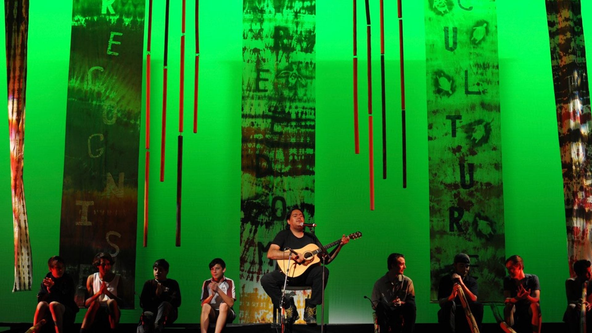 Person sitting on a chair with a microphone, playing guitar. 8 young people with didgeridoos sit around them