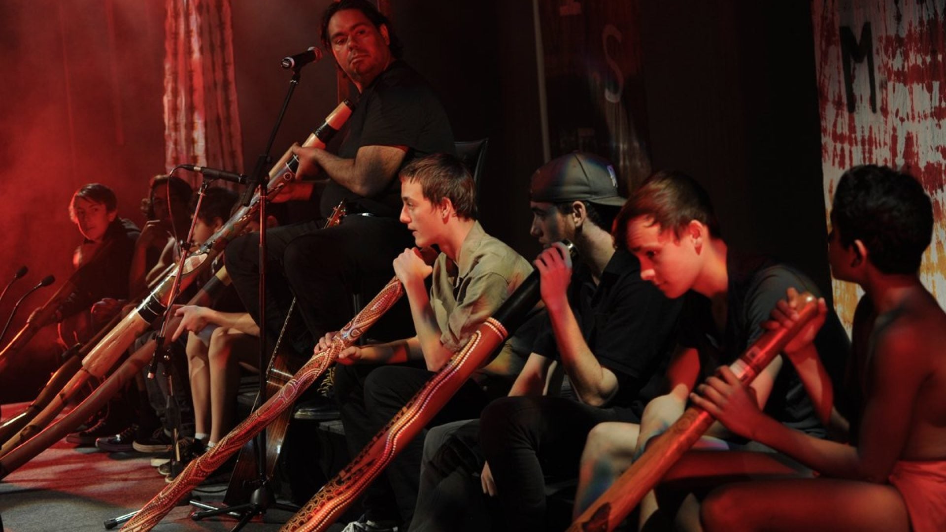 Person sitting on a chair with a microphone, playing didgeridoo. 8 young people with didgeridoos sit around them