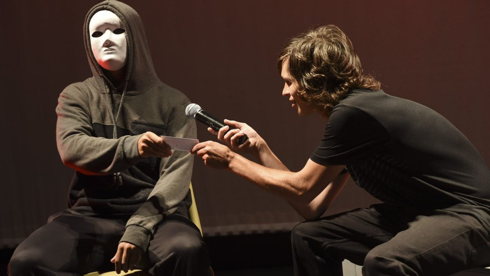 Two people sitting on chairs on a stage. One person is wearing a while mask
